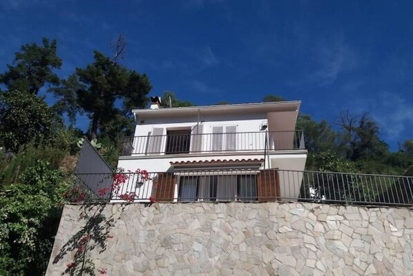 A two-story building with a stone facade, a balcony with metal railings, and a clear blue sky in the background. - Between Land and Sea - A Quiet House in Rosamar (Canyet de Mar)