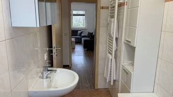 Een moderne badkamer met een wastafel, bad en handdoekhouder. Een gang leidt naar een woonruimte met een bank en een raam.