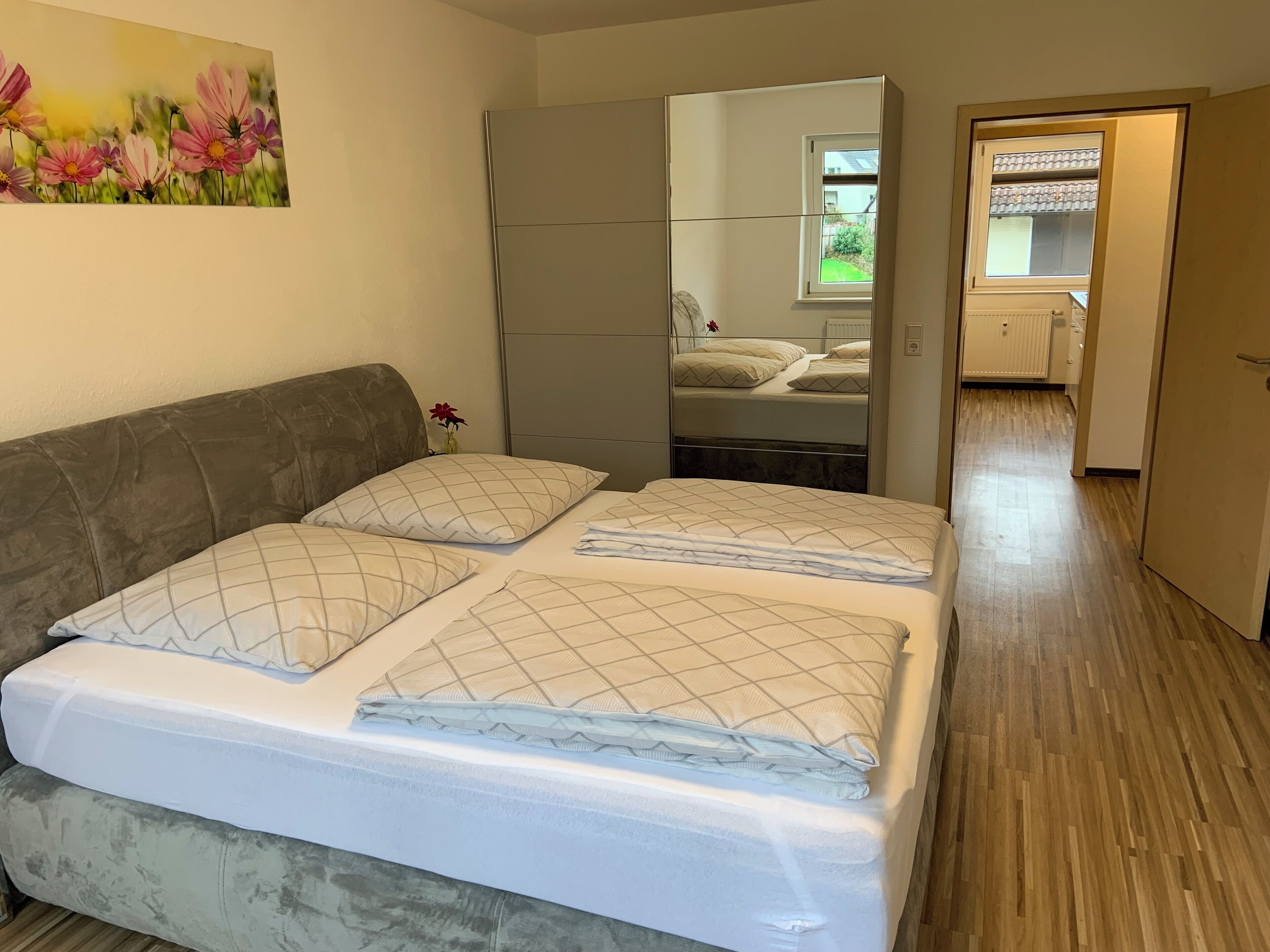 Ein Schlafzimmer mit einem Bett, einem grauen Sofa, einem Bild an der Wand und Blick auf die Küche durch eine offene Tür.