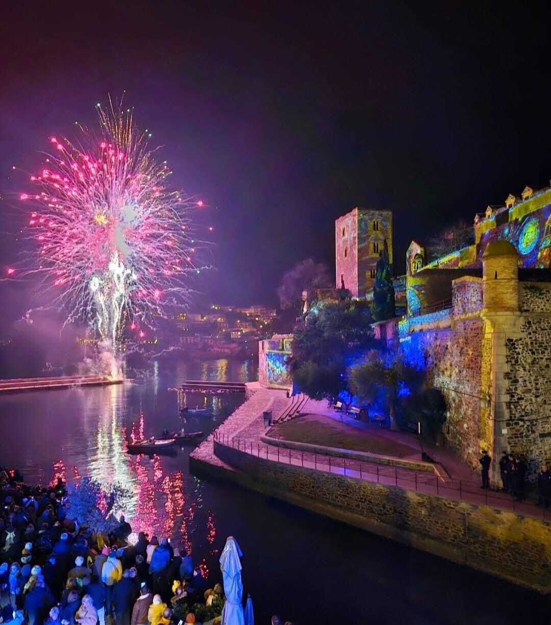 Un pueblo histórico con una estructura similar a un castillo, un muelle y una multitud de personas observando fuegos artificiales.