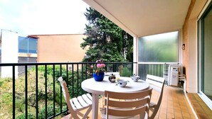 Un balcón con una mesa redonda y dos sillas, una planta en maceta con flores rosadas, y vistas a un árbol y un edificio al fondo.