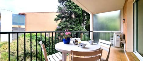 Un balcón con una mesa redonda y dos sillas, una planta en maceta con flores rosadas, y vistas a un árbol y un edificio al fondo.