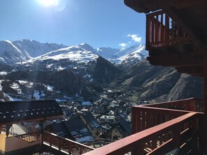 A mountain town with snow-capped peaks in the background, a view from a balcony with wooden railings, and a small village below.