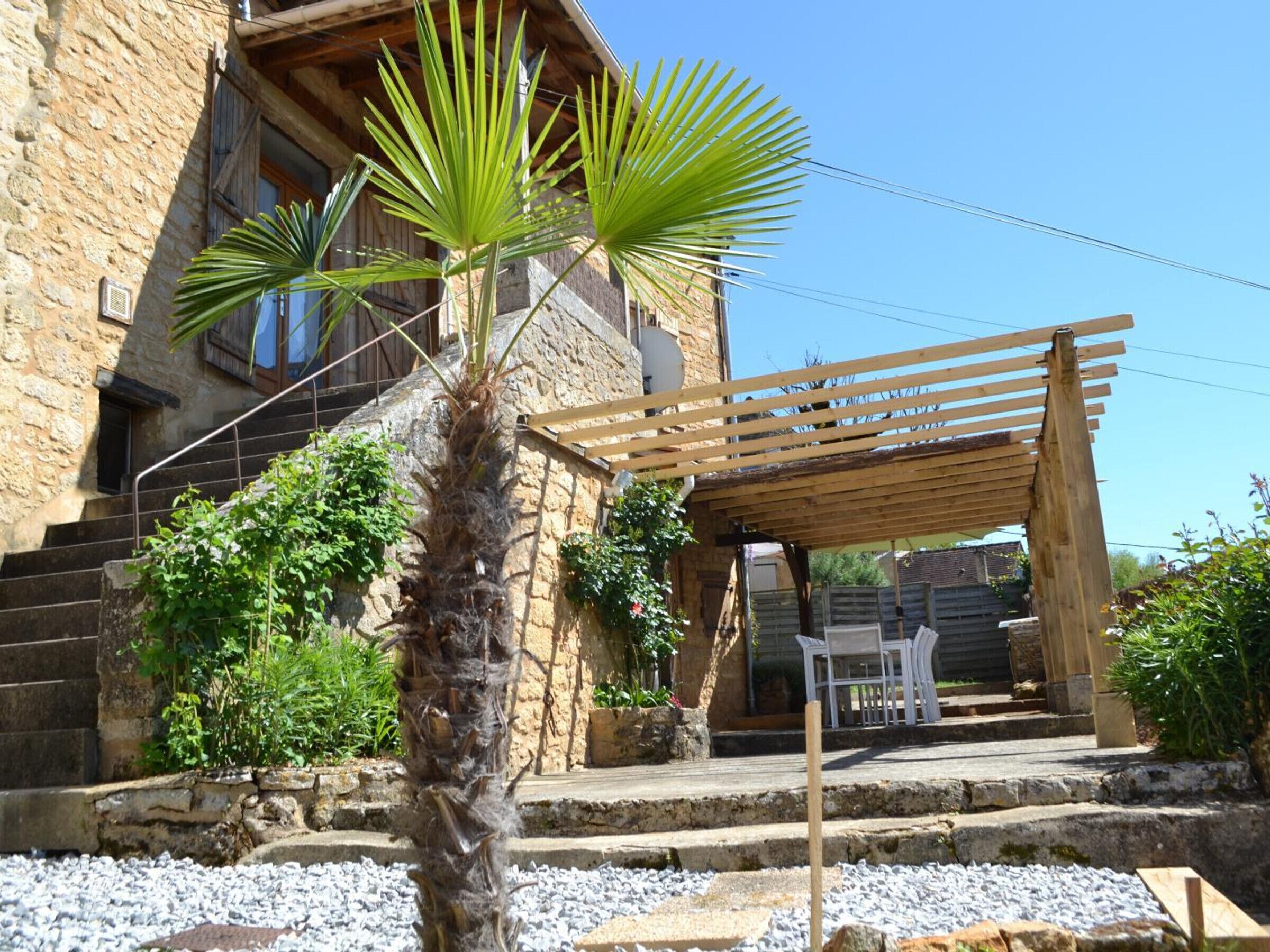 Un bâtiment en pierre avec une pergola en bois et un palmier au premier plan.