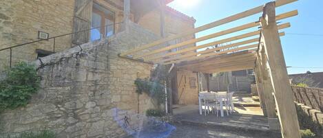 Edificio de piedra con una pérgola de madera y un patio con muebles de exterior.