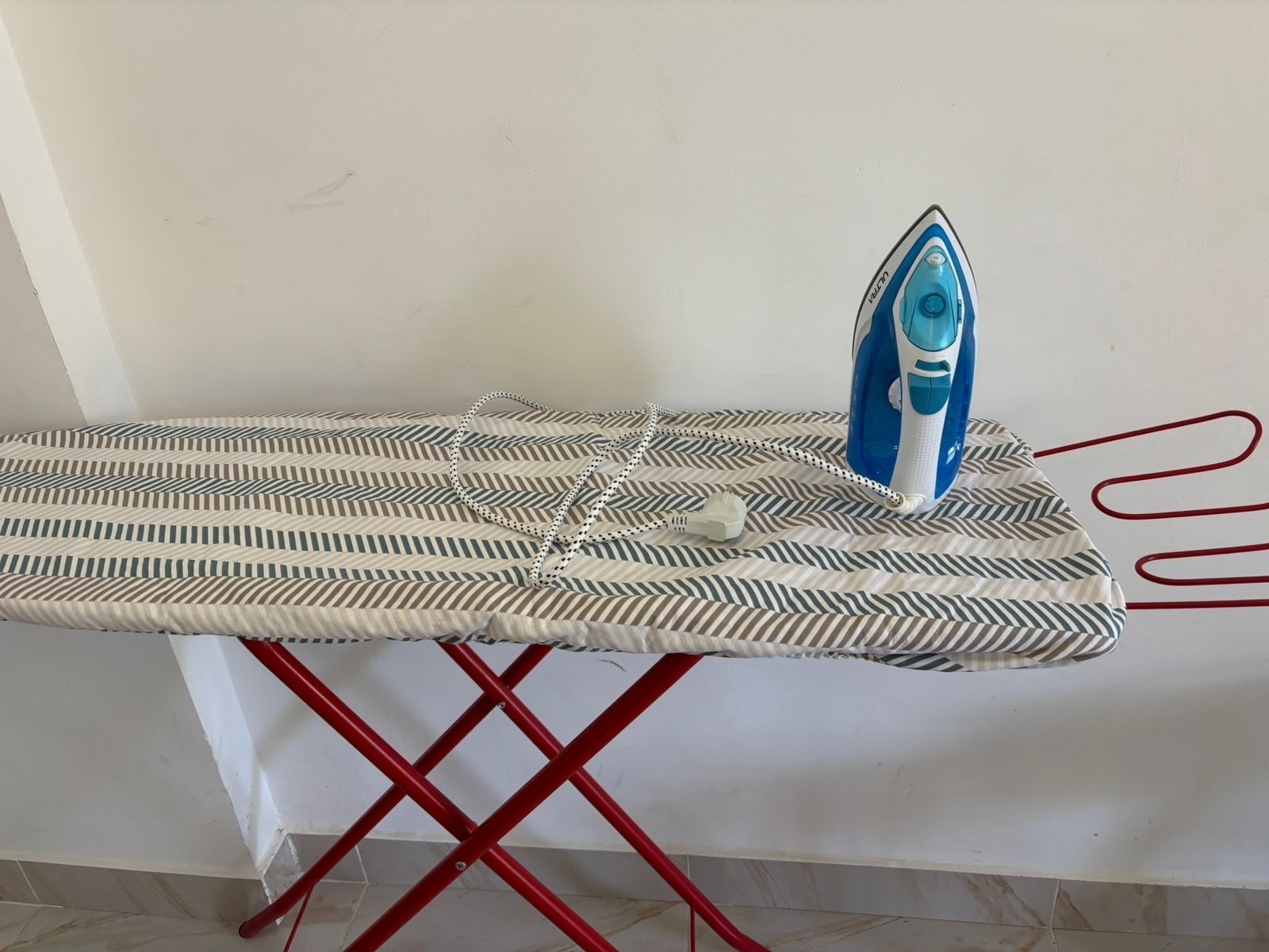 Une planche à repasser recouverte d’un tissu à rayures, un fer à repasser bleu et un support rouge.