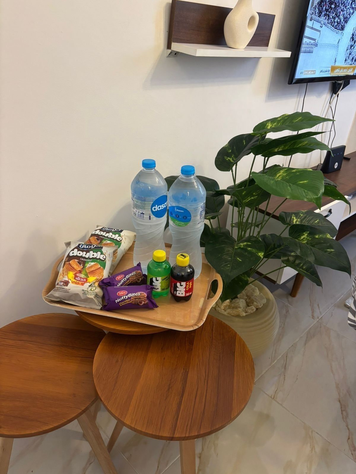A tray with snacks, water bottles, and a bottle of sauce on a wooden table in a room with a plant and a TV on the wall.