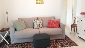 En stue med en sofa, mønstrede puder, et tæppe, en lille skammel, et hvidt skab og et billede på væggen.