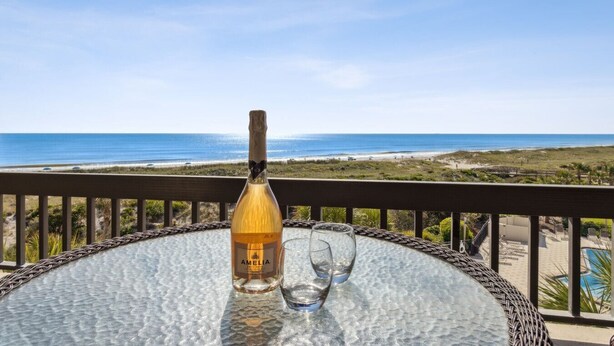 Een fles Amelina Brut op een glazen tafel, met twee glazen, met uitzicht op een strand.