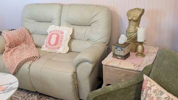 Une pièce avec un canapé beige, un fauteuil vert, une petite table ornée d’objets décoratifs, un miroir et un tapis à motifs.