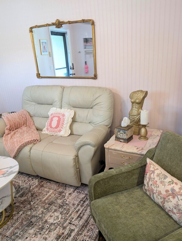 Una habitación con un sofá color crema, un sillón verde, una mesita con adornos, un espejo y una alfombra estampada.