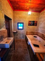 A bathroom with a stone wall, a wooden door, a window, a painting, a sink, and a bathtub.