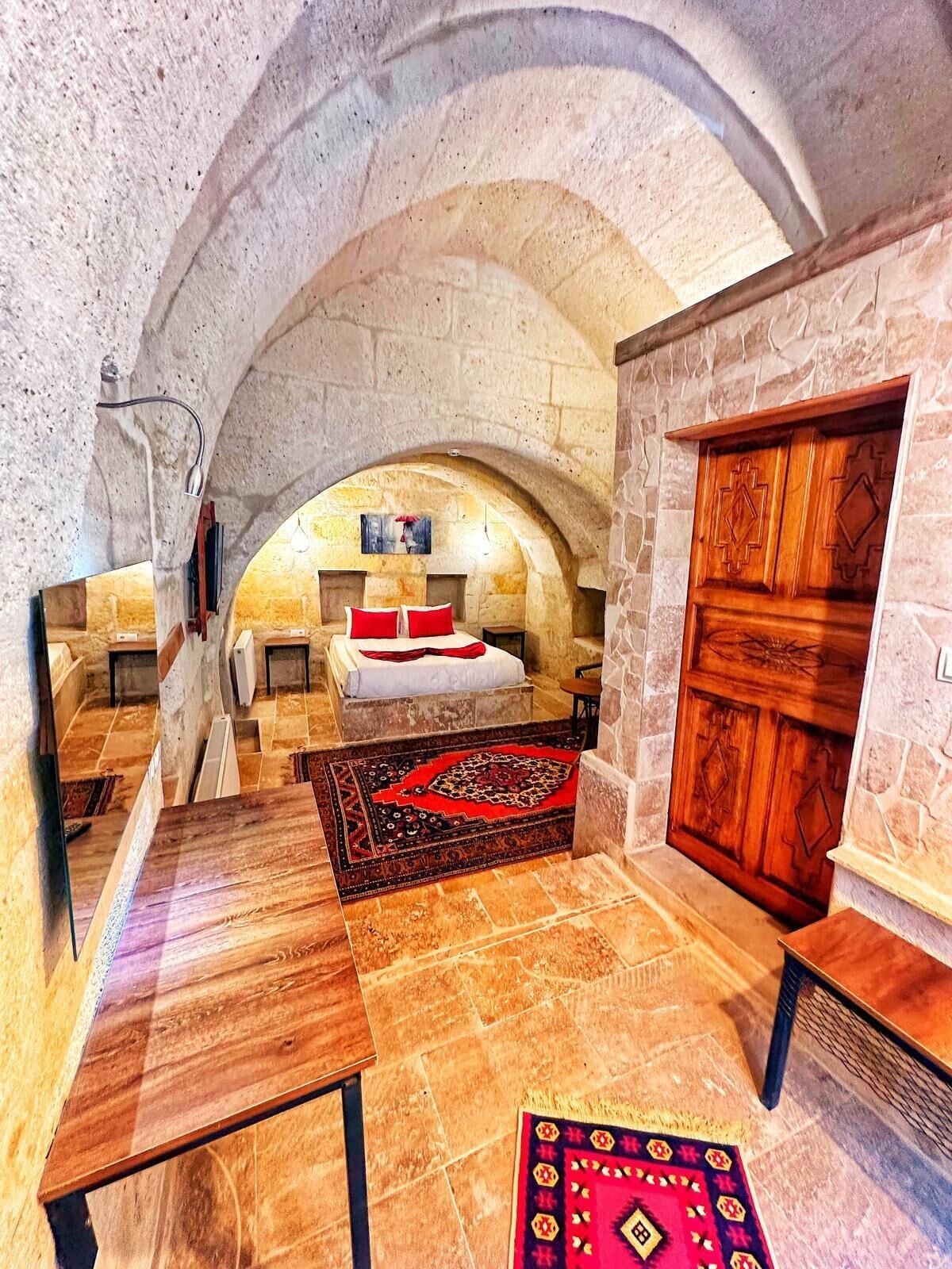 A stone-walled room with a bed, a wooden wardrobe, and a red patterned rug.