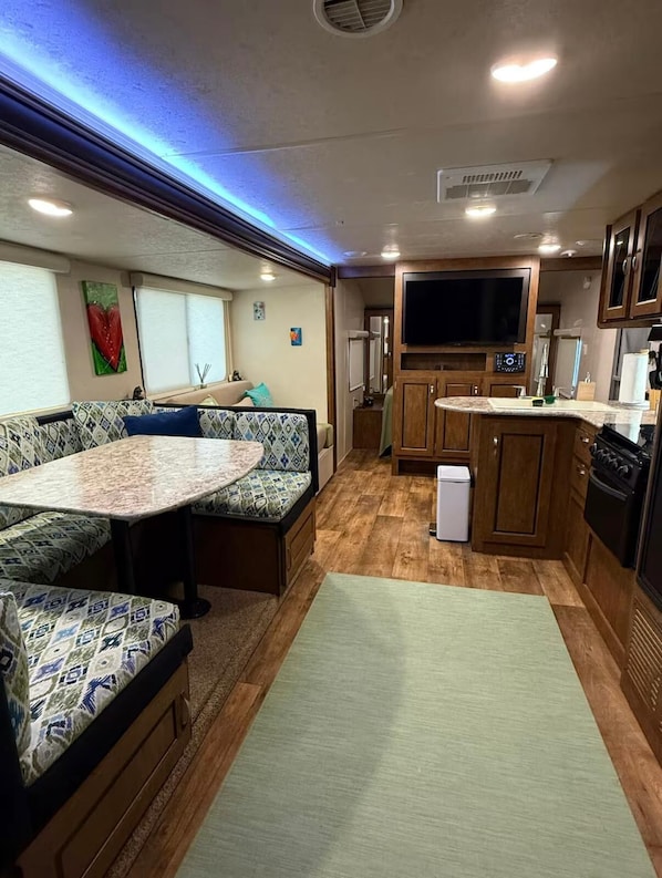 A compact RV interior with a kitchenette, a dining area with a round table, and a seating area with patterned cushions.