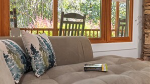 Un canapé beige agrémenté de deux coussins à motifs et d’un livre, placé devant une fenêtre à cadre en bois, qui offre une vue sur des arbres et une terrasse en bois.