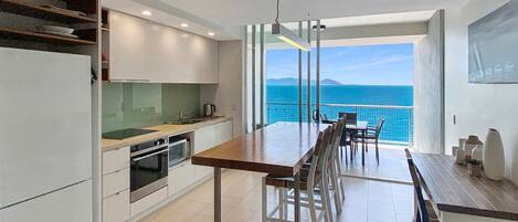 A modern kitchen with a dining area, large windows, and a view of the sea.