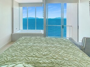 A bedroom with a large bed, a window with a view of the sea, and a bathtub.