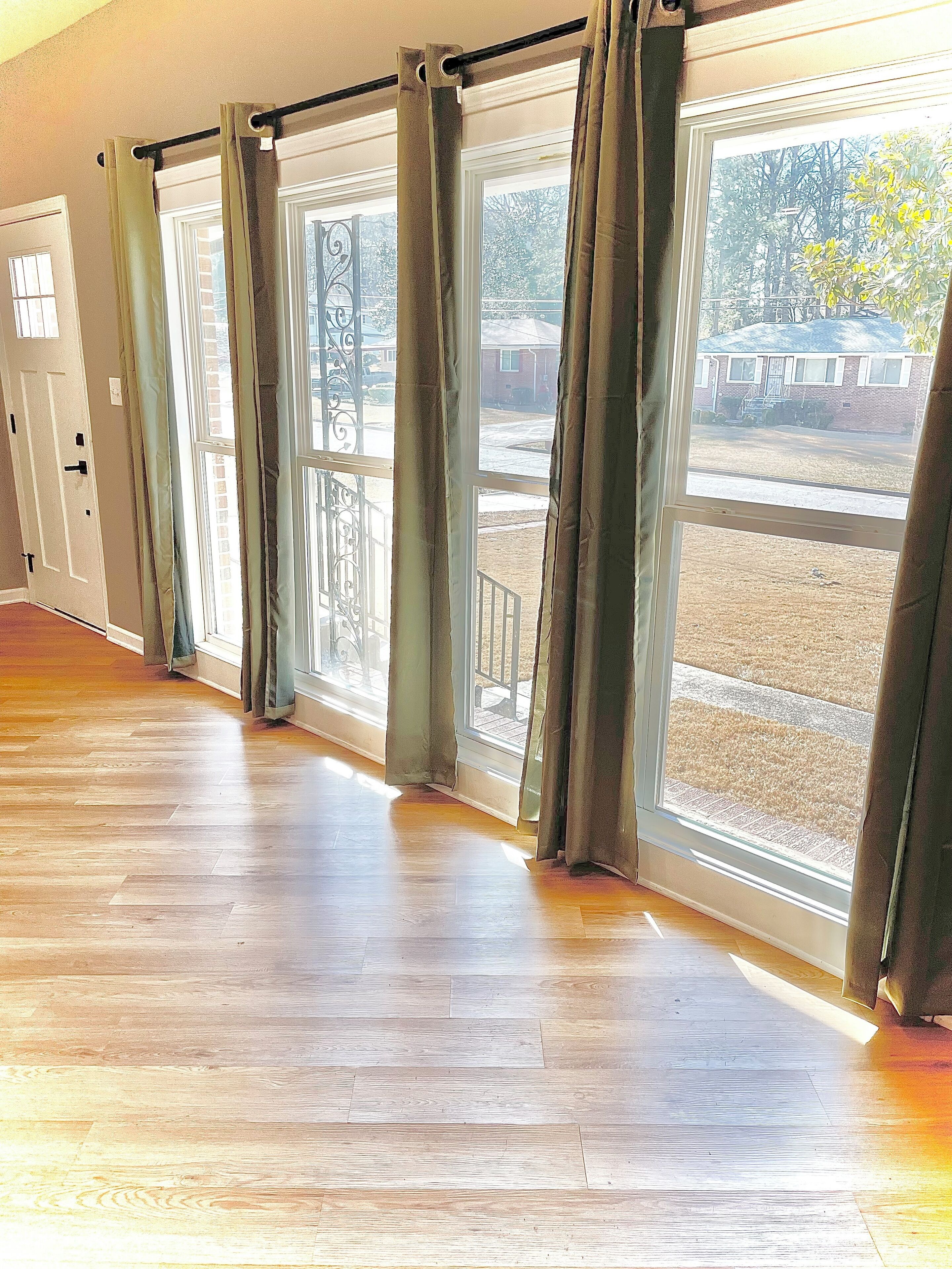 A room with large windows, wooden flooring, and a view of a residential area.