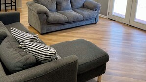 A modern living room with a grey sofa, a matching ottoman, and a houndstooth pillow. A kitchen with white cabinets and a dining area with a table and chairs are visible in the background. - Lovely 3-bedroom house with AC in vibrant Atlanta (Atlanta)