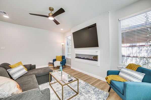 A modern living room with a sectional sofa, a blue armchair, a coffee table, a flat-screen TV mounted on the wall, and a fireplace below it.