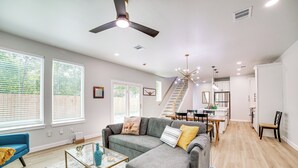 A spacious living room with a gray sectional sofa, a glass coffee table, and a blue armchair. A staircase leads to the upper floor, and the room has large windows with blinds.