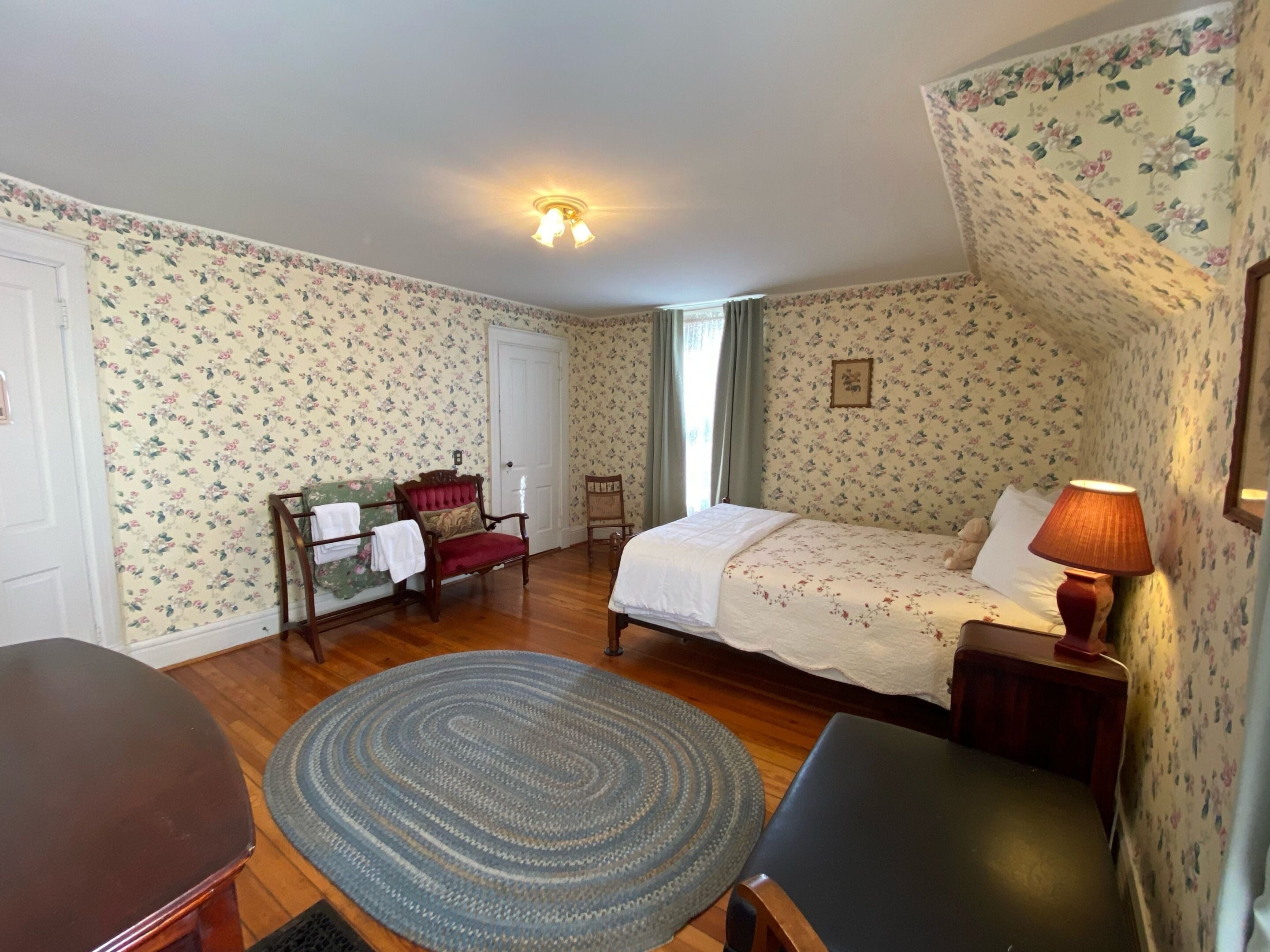 A bedroom with floral wallpaper, a bed with white linens, a wooden floor, a desk, a chair, and a lamp.