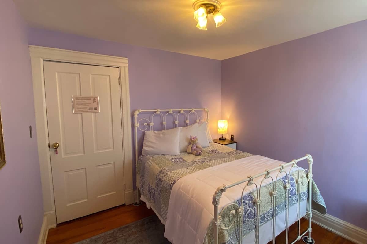 A bedroom with a white door, a bed with a metal frame, a nightstand with a lamp, and a purple wall.