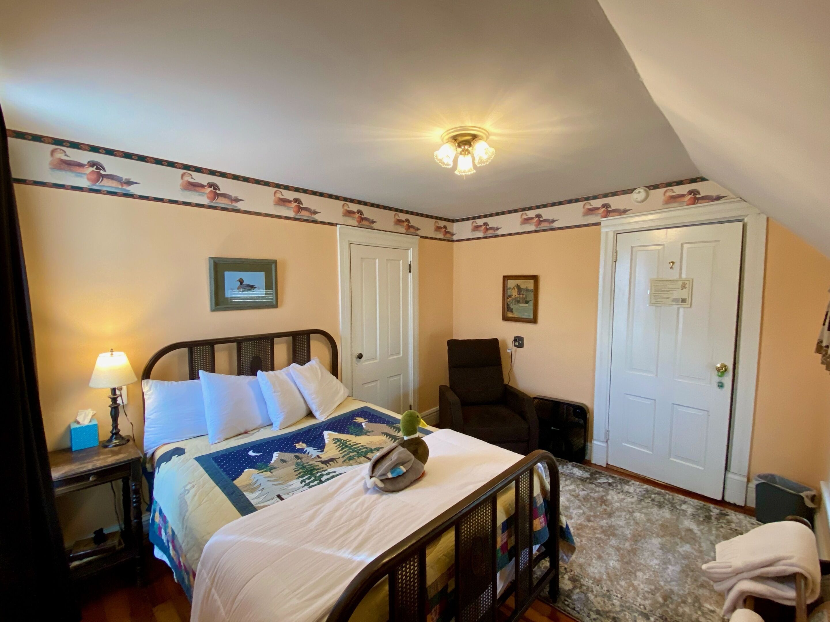 Habitación de hotel con cama, mesita de noche, lámpara, silla y dos puertas.