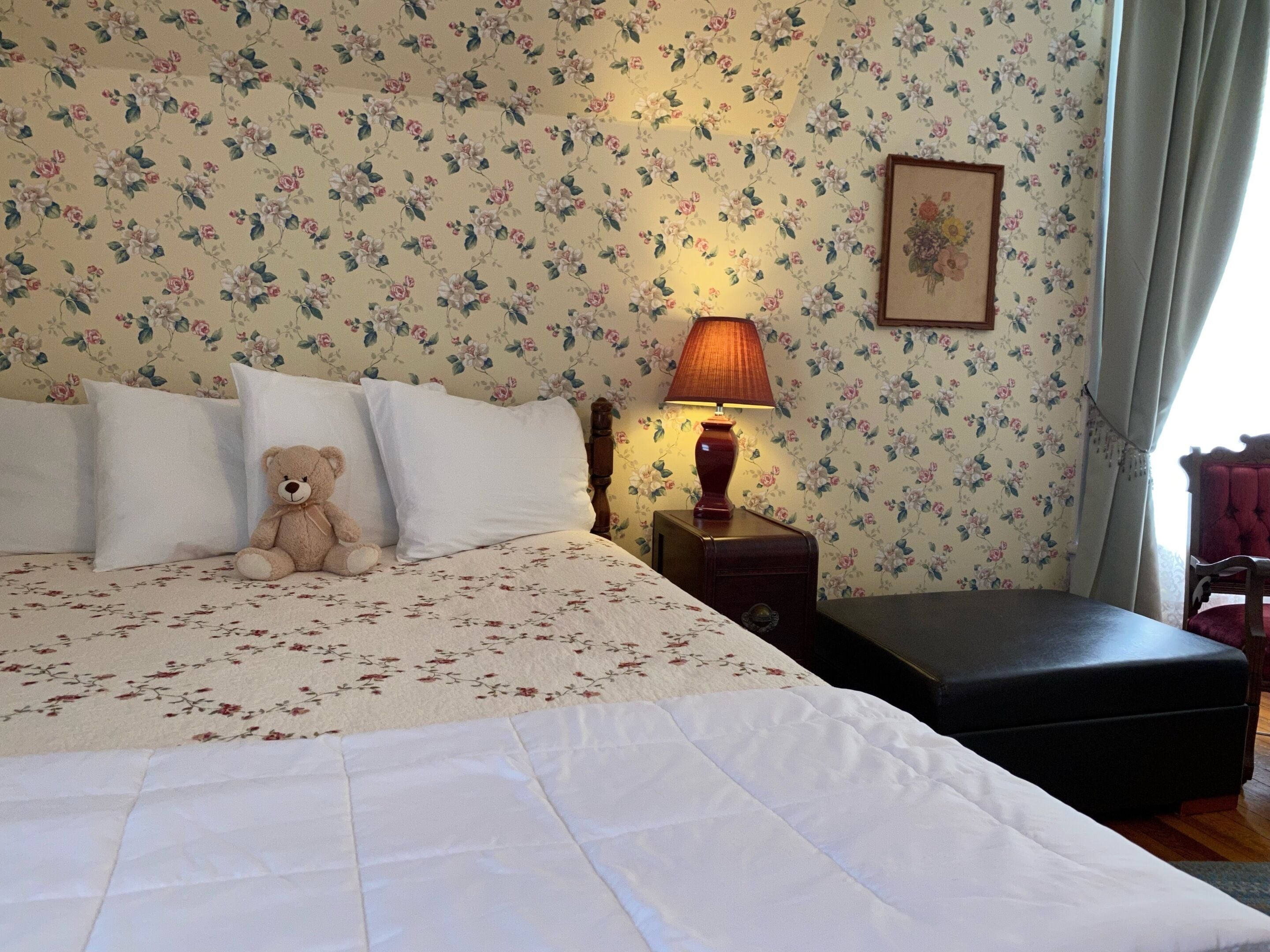 A neatly made bed with white linens and a teddy bear on the pillows, a bedside lamp, and a floral wallpaper.