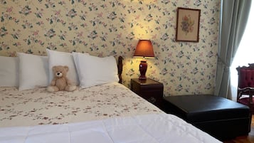A neatly made bed with white linens and a teddy bear on the pillows, a bedside lamp, and a floral wallpaper.