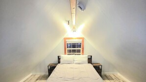A bedroom with a wooden floor, a bed with white bedding, a nightstand, a small table, and a window with blinds.