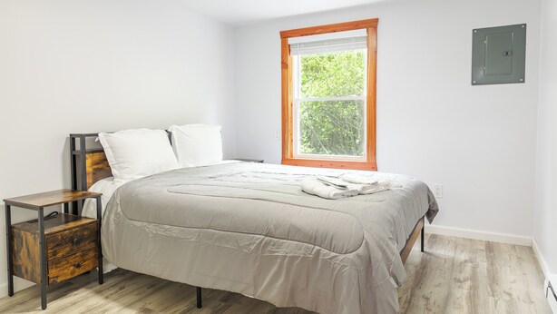 A bedroom with a large bed, a nightstand, a window with blinds, and a wall-mounted light switch.