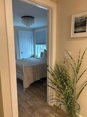 Un pasillo estrecho que conduce a un dormitorio con una cama, una ventana con persianas, una puerta y una planta en maceta.