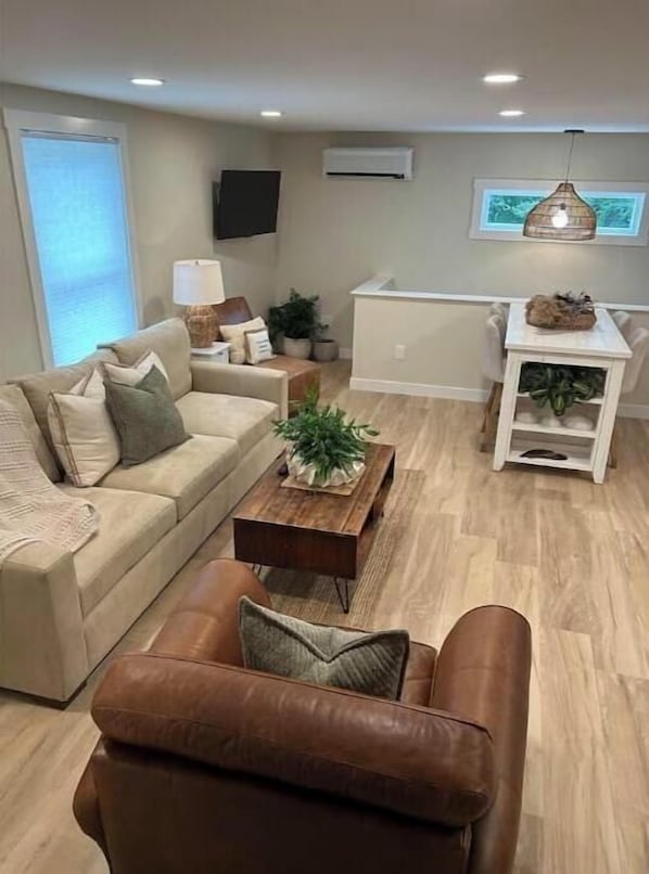 Una sala de estar con un sofá, una mesa de centro, un sillón de cuero marrón, una mesita blanca con una planta y un televisor montado en la pared.