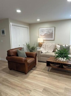 Una sala de estar con un sillón de cuero marrón, un sofá color beige, una mesa de centro de madera y una puerta blanca.