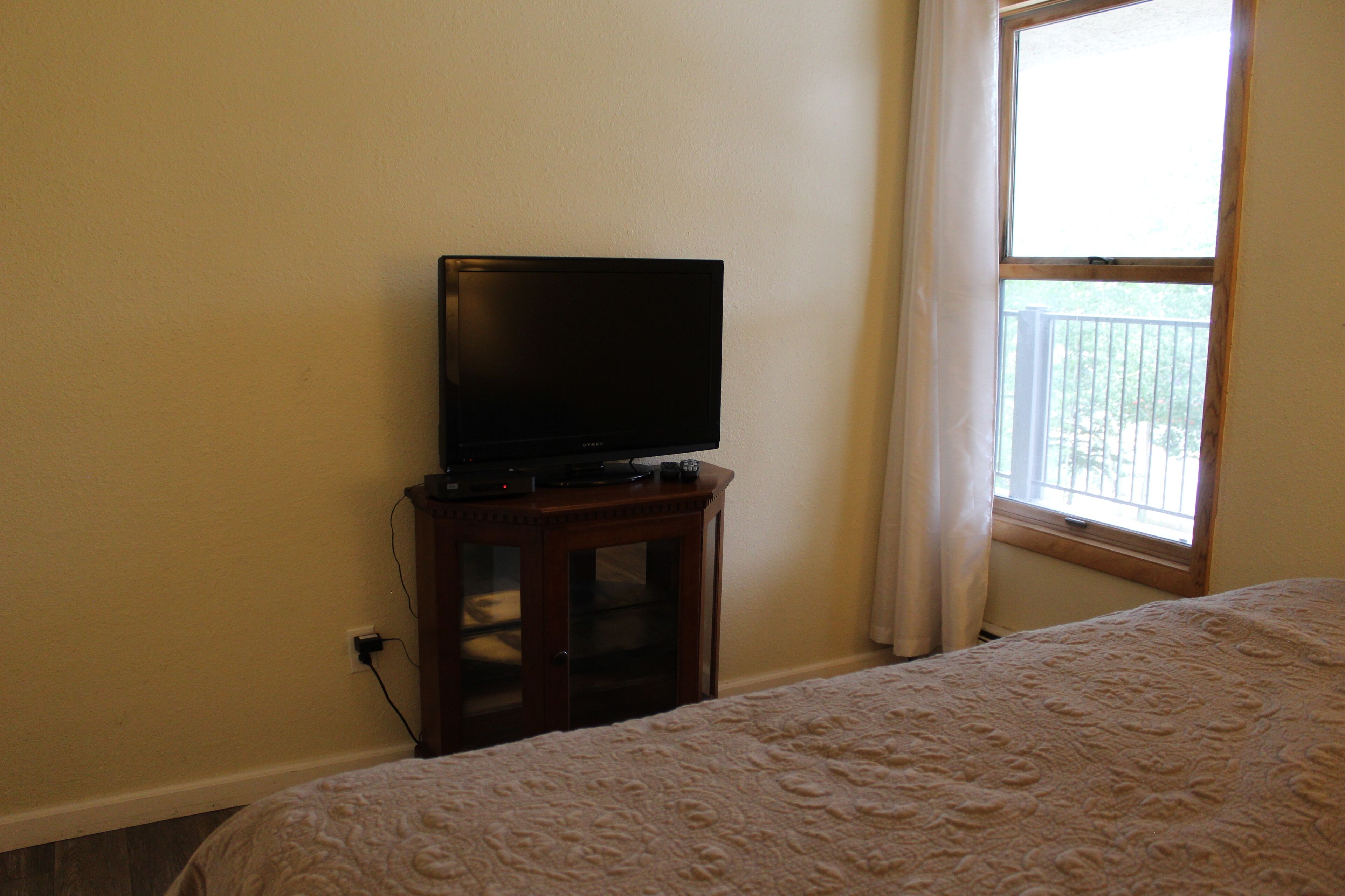 A bedroom with a bed, a television on a wooden stand, and a window with a view of a balcony.