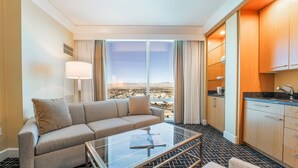 A hotel room with a sofa, a glass coffee table, a view of a desert landscape through large windows, and a kitchen area with wooden cabinets and a black countertop.