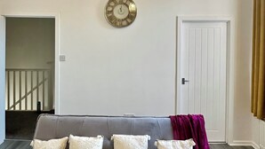 Uma sala de estar com sofá cinza, almofadas decorativas, um candelabro, um relógio de parede e uma porta.