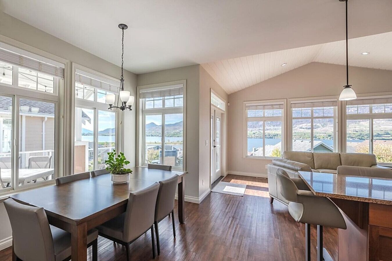 A spacious dining area with a long table and chairs, large windows with a view, and a kitchen area with a countertop and seating.