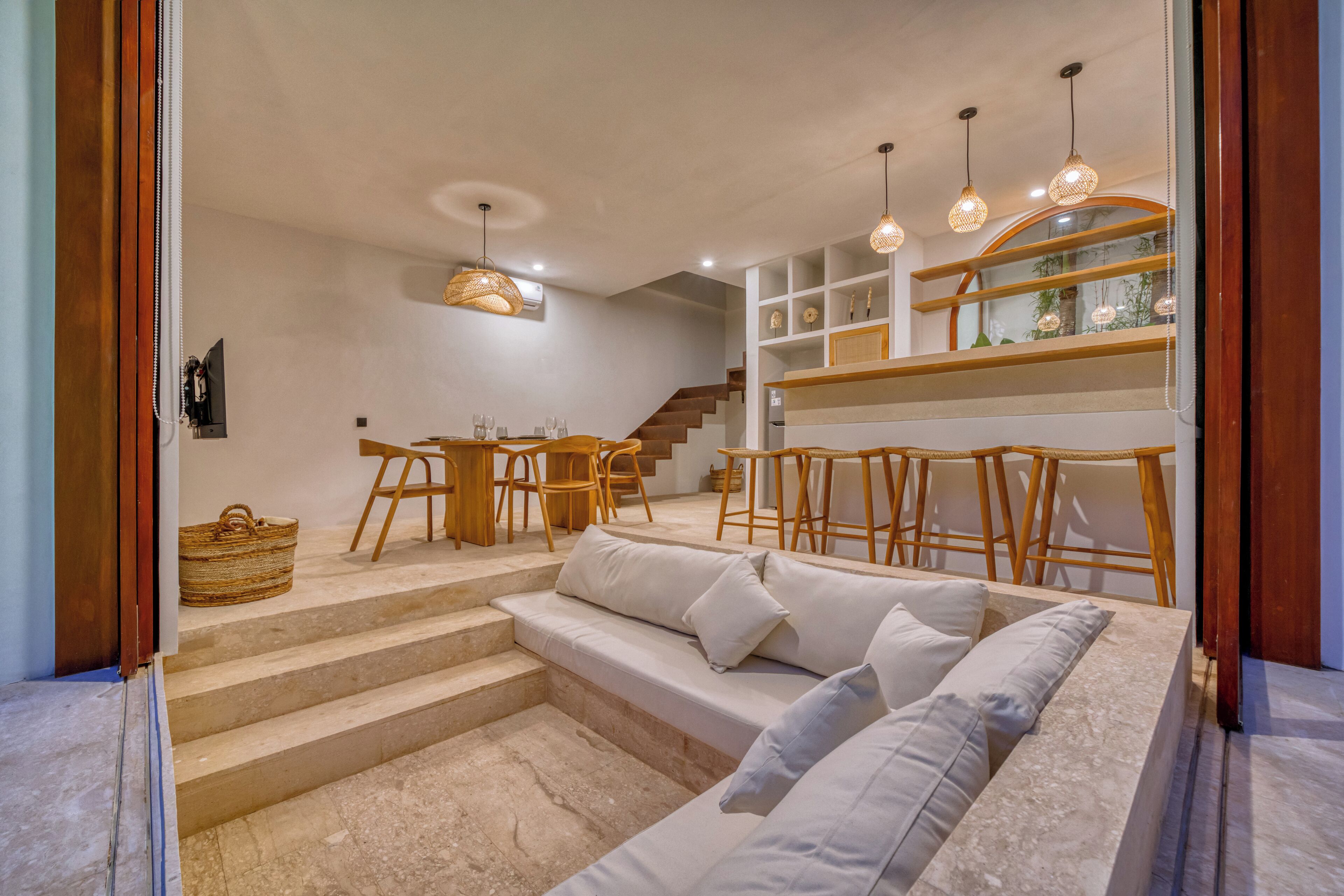 A modern living space with a sofa, dining table, and chairs. There are steps leading to a staircase and a shelf with decorative items.