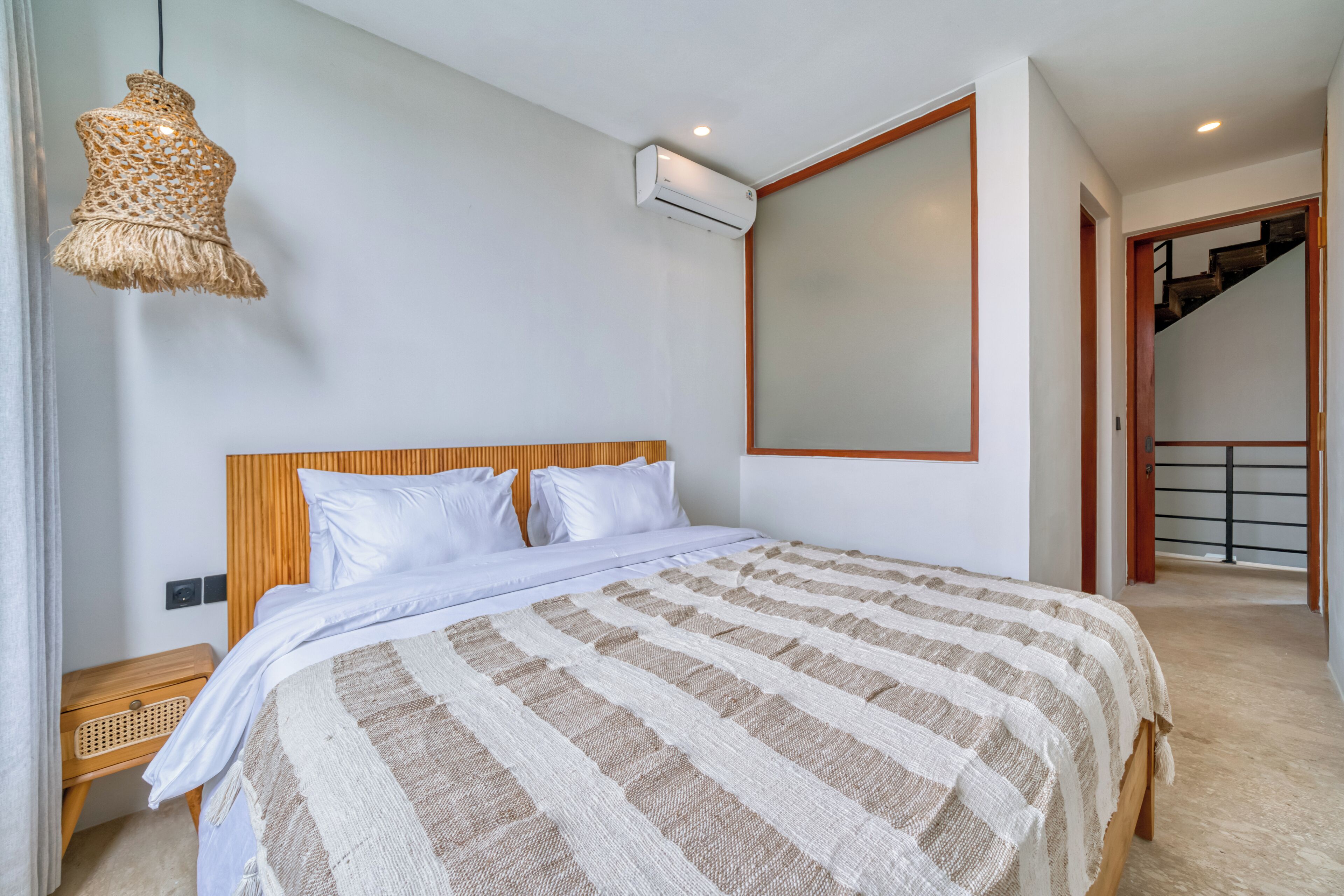 A bedroom with a bed, a hanging light fixture, an air conditioning unit, and a view of a staircase.