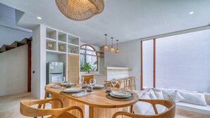 A modern dining area with a wooden table set for six, surrounded by wooden chairs. A built-in shelving unit is visible to the left, and a staircase leads to an upper level. - Villa Gloria Canggu (Canggu)