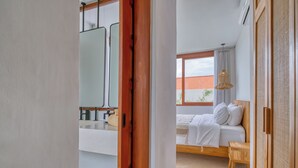 A bedroom with a bed, a wooden chair, a window, and a bathroom with a mirror. - Villa Gloria Canggu (Canggu)
