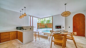 A modern kitchen with wooden cabinets, a dining area with a round table and chairs, a built-in oven, and a large window with a view of greenery. - Villa Gloria Canggu (Canggu)