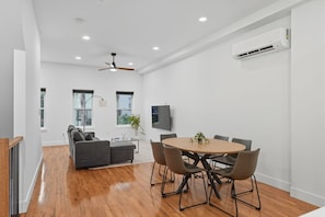 A modern living room with a wooden floor, a sofa, a dining table with chairs, a ceiling fan, and an air conditioning unit.