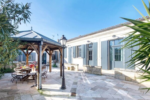 A covered outdoor dining area with a table and chairs, a pergola, and a building with large windows and shutters.
