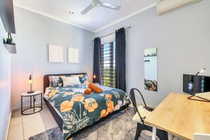 Quarto com cama, escrivaninha, cadeira, ventilador de teto, janela com cortinas e TV na parede.