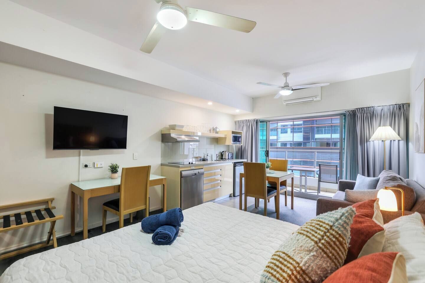 A hotel room with a bed, a desk with a chair, a television, a kitchenette, and a balcony with curtains.