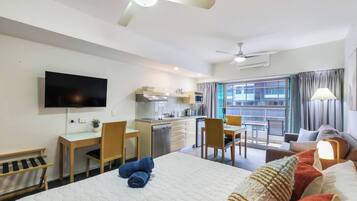 A hotel room with a bed, a desk with a chair, a television, a kitchenette, and a balcony with curtains.