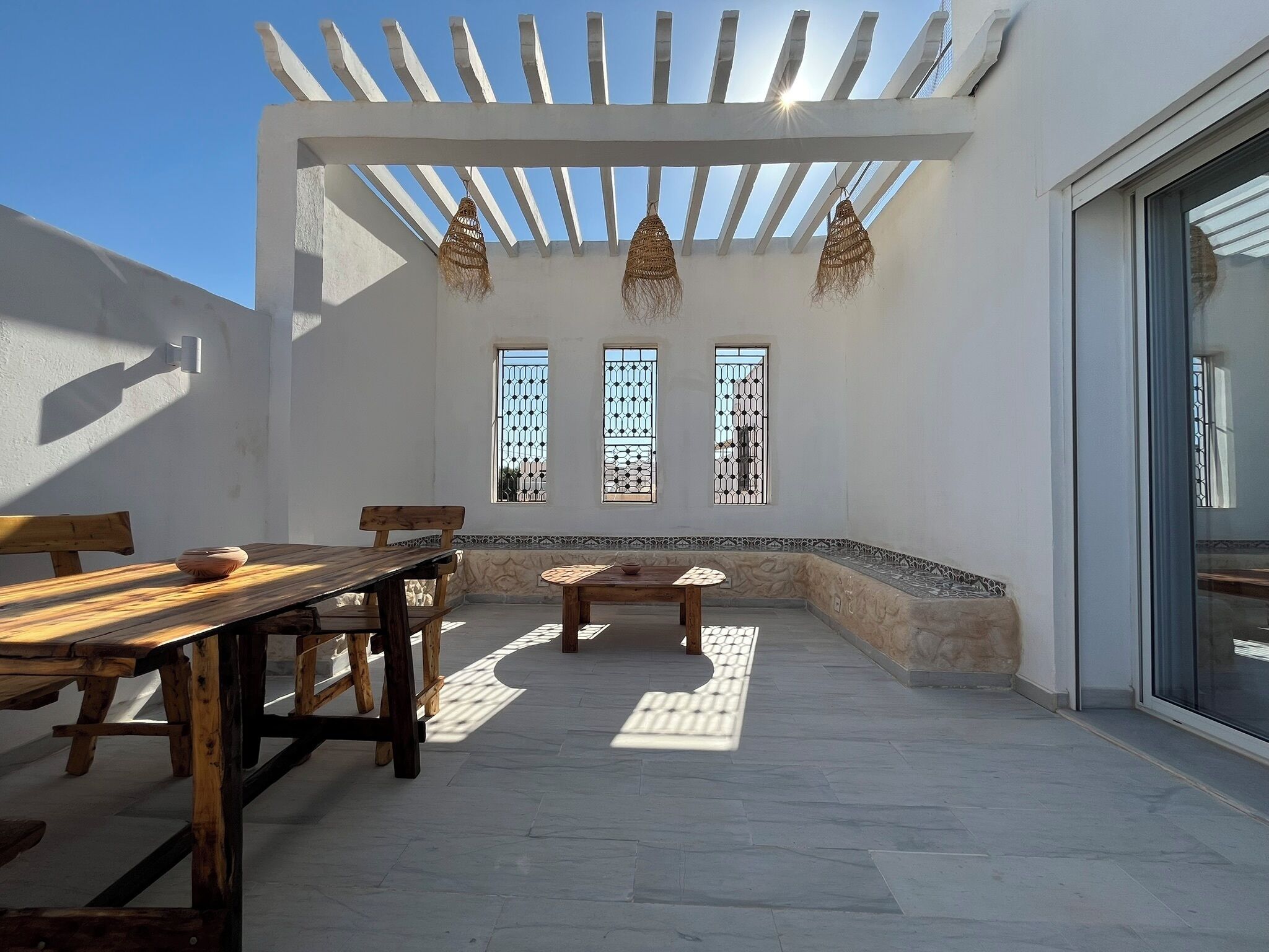 An outdoor patio with a wooden table and benches, a pergola with hanging lanterns, and three windows with decorative grills.